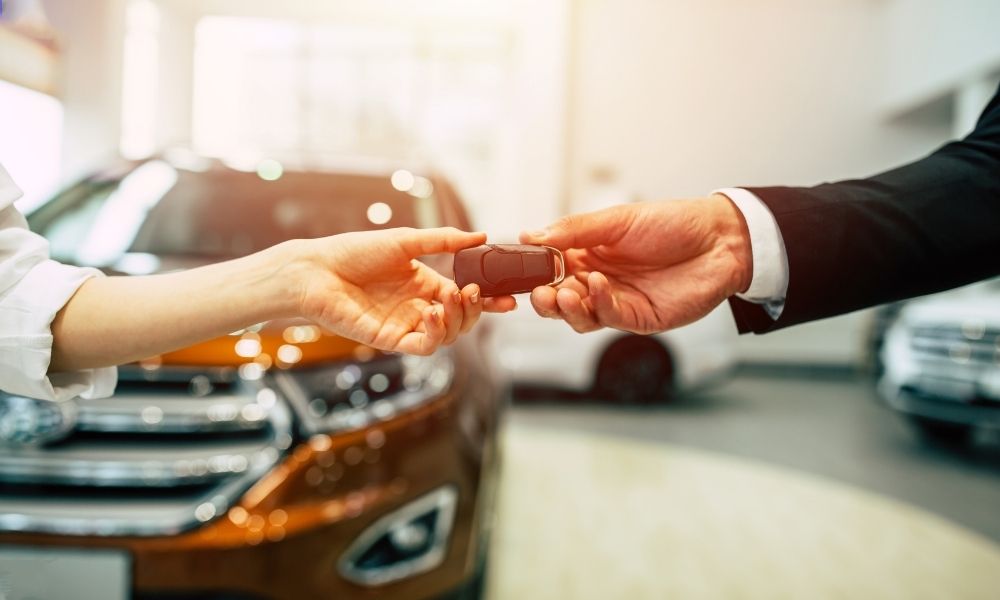 A man in a suit handing keys over to a person with an outstreched hand in a car.