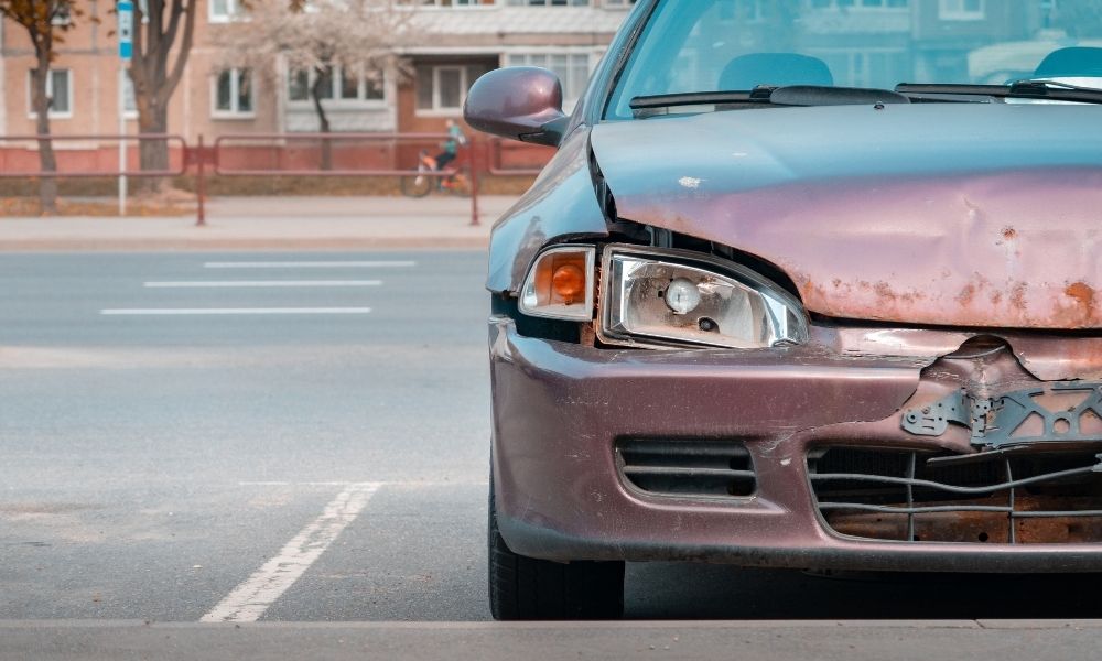 A maroon colored car with a busted headlight from an accident.